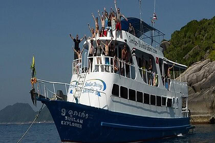 the liveaboard Similan Explorer packed with divers waving from the top deck, anchored by a rocky island
