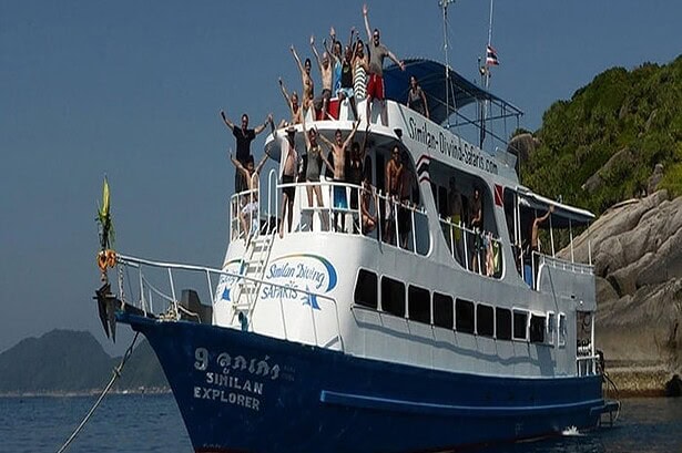 the liveaboard Similan Explorer packed with divers waving from the top deck, anchored by a rocky island