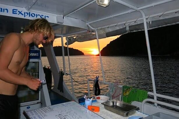 Crew member near the dive briefing whiteboard and drinks fridge on the Similan Explorer's rear deck at sunset