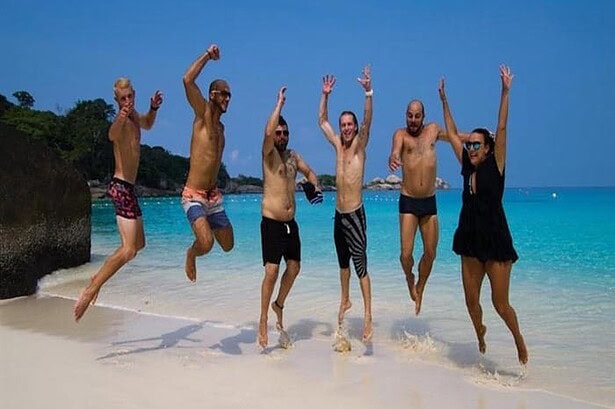 Six divers jumping for a group photo on a white sand beach with turquoise water