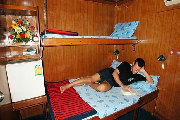 Diver relaxing with a book in a wood panelled twin bunk cabin aboard the MV Marco Polo liveaboard
