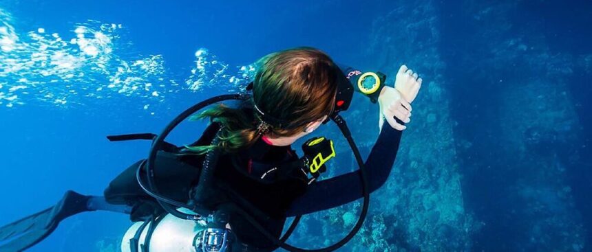 Diver in a wetsuit checking a wrist-mounted dive computer while hovering beside a reef wall in blue water
