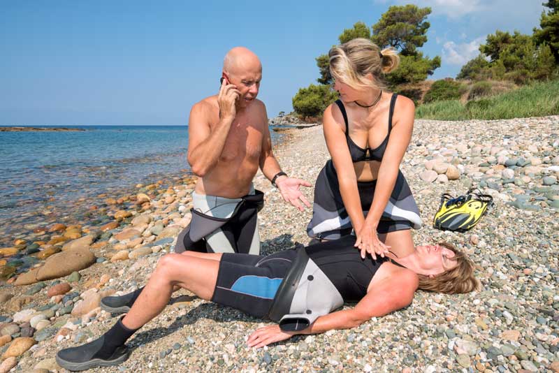 Two people give first aid to an unresponsive diver on a rocky beach while one calls for help