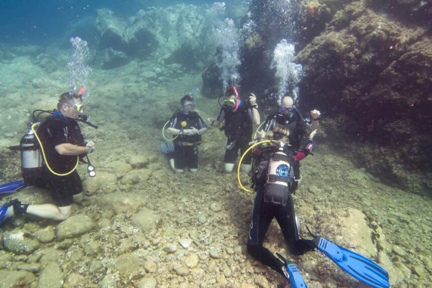 Five scuba divers kneeling on a rocky seabed exhaling bubbles during a guided dive