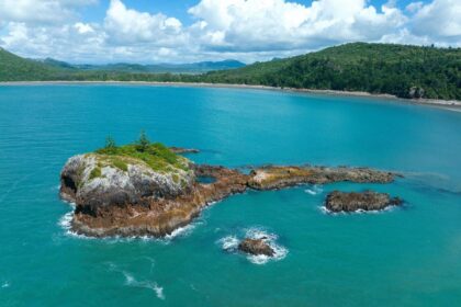 Aerial view of a rocky islet in a turquoise bay backed by forested headlands in Australia