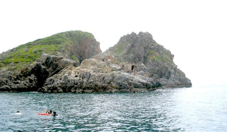 Rocky islet with a small hut rising from calm sea at Madonna Rock in Vietnam, snorkelers in the water