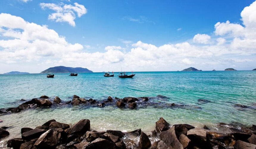 Wooden fishing boats anchored off a rocky shore in turquoise water with islands on the horizon in Vietnam