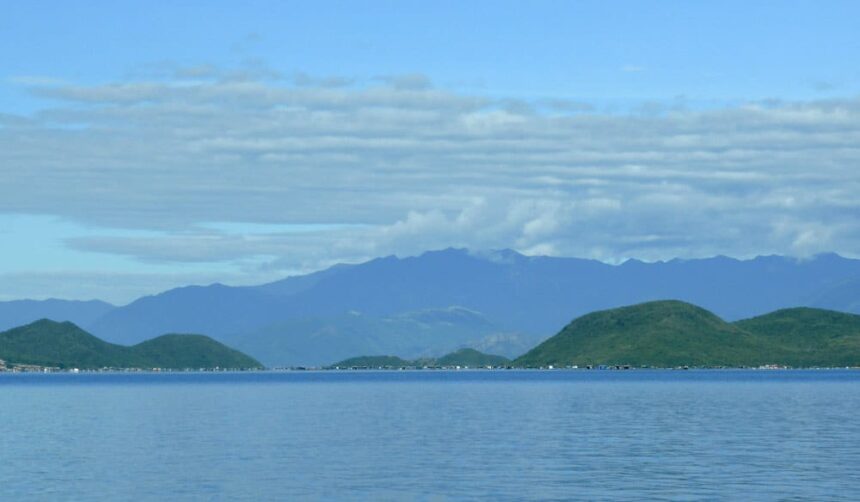 Calm blue sea backed by forested green hills and a distant floating village in Vietnam