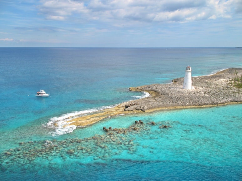 A white lighthouse stands on a rocky point above clear turquoise reef water in the Bahamas