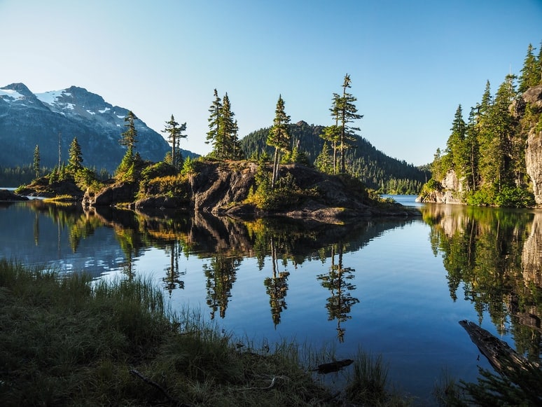 A calm forested lake reflects snow capped peaks on Vancouver Island