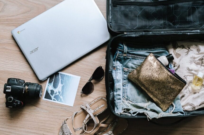 An open suitcase packed with clothes next to a laptop, camera and sunglasses