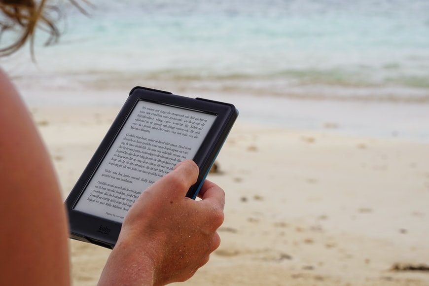 A hand holds a Kobo e-reader on a sandy beach with turquoise water beyond