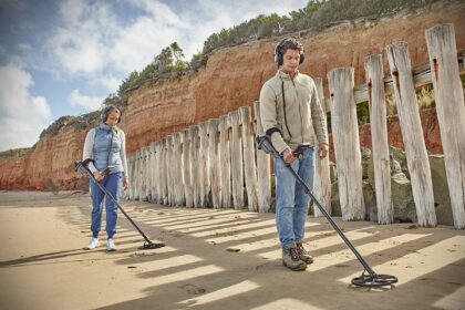 Two detectorists in headphones sweeping metal detectors along a sandy beach beside weathered wooden posts
