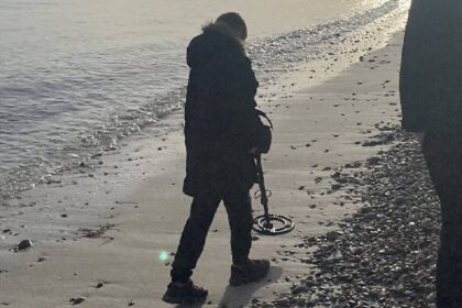 Detectorist in a winter coat sweeping a metal detector along a pebbly shoreline in low backlit sun