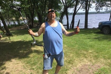 Man standing on grass by a lake holding a metal detector in one hand and a digging tool in the other