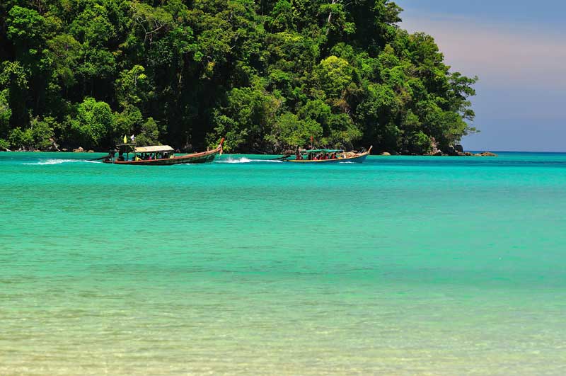 Longtail boats anchored in turquoise water beneath jungle-covered cliffs on a Thailand coastline