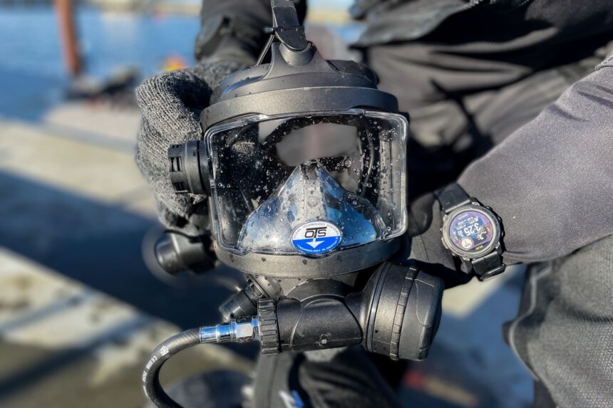 ots guardian mask product closeup Gloved hand holding the OTS Guardian full face mask with its regulator attached, at the water's edge