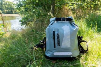 IDRYBAG waterproof roll-top backpack standing in long grass by the water, beaded with droplets