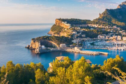 Golden evening light over a Mallorca harbour town, with boats moored below pine-covered cliffs
