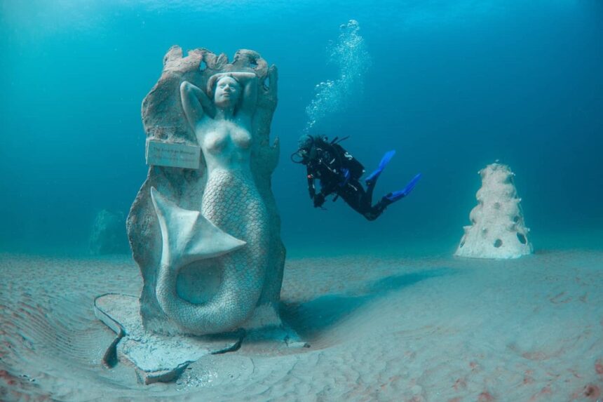 Scuba diver swimming past a mermaid statue on the sand at an artificial reef