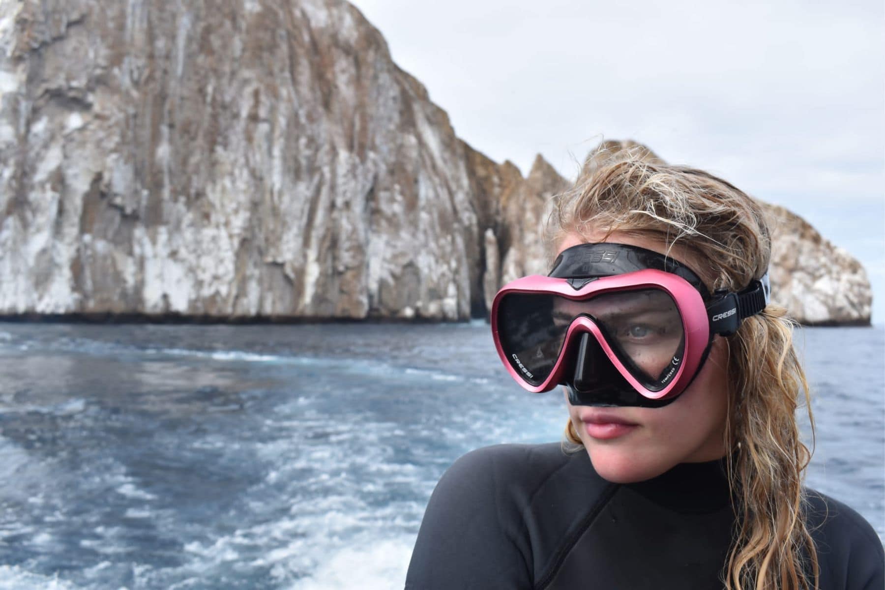 Our reviewer on a dive boat wearing the pink Cressi A1 mask, rock formation behind her