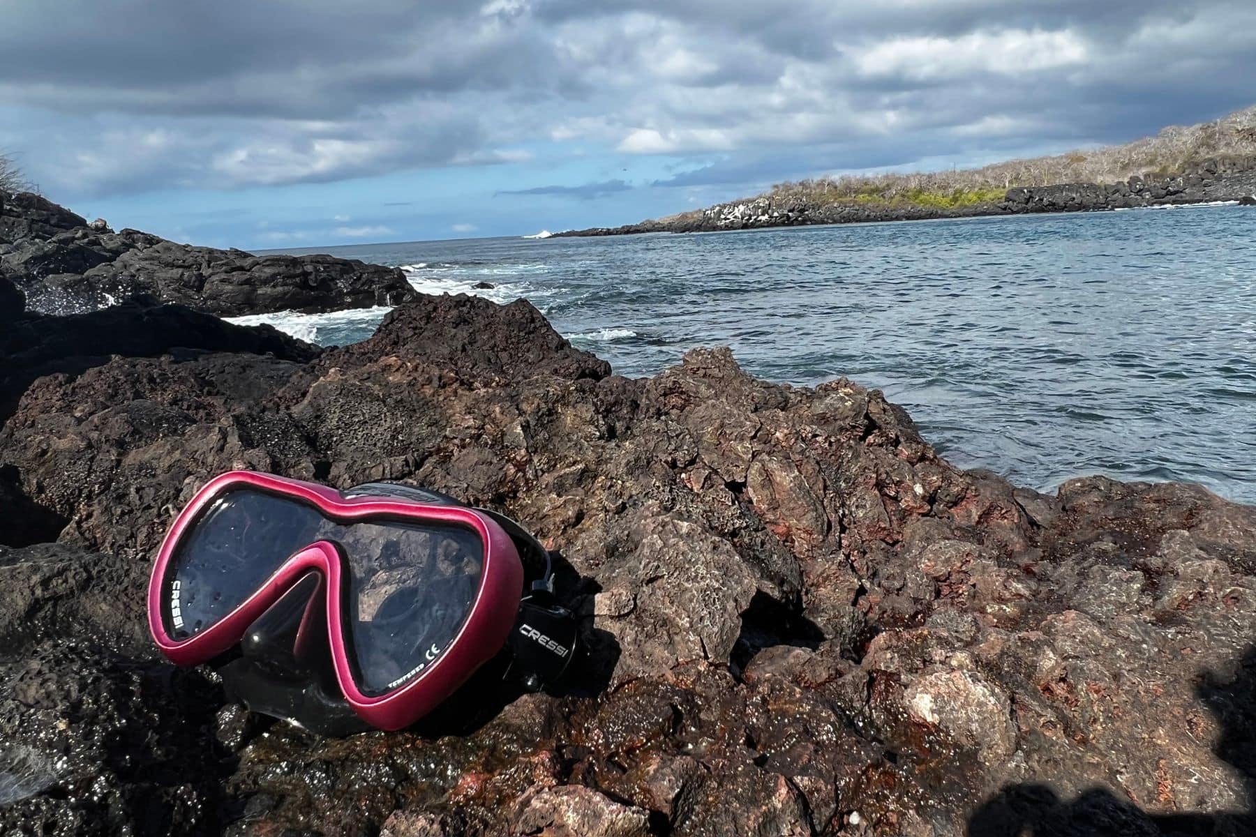 Pink and black Cressi A1 dive mask resting on volcanic rock beside a rocky coastline
