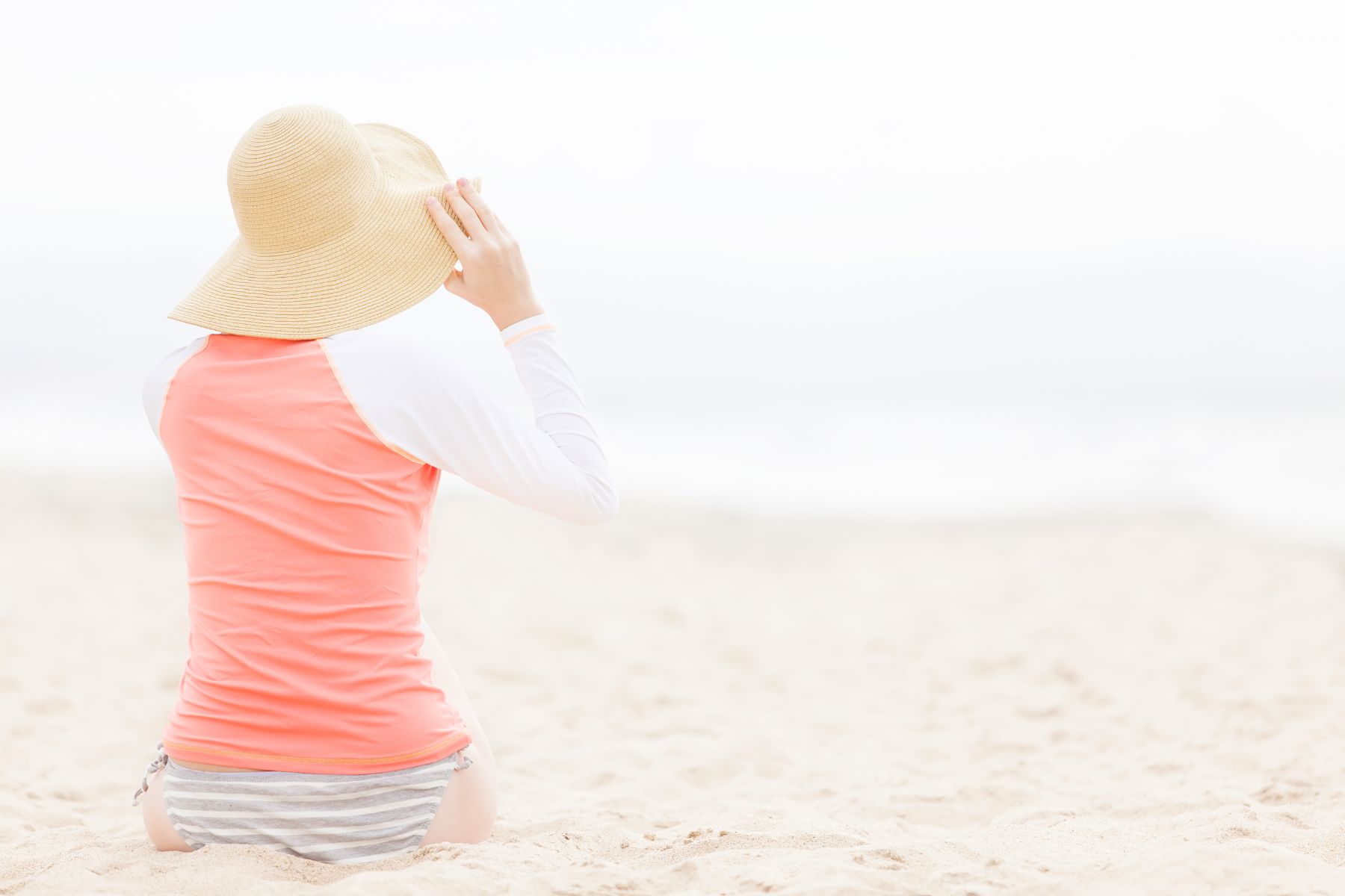 Woman in a coral and white long-sleeve rash guard and straw sun hat sitting on a beach