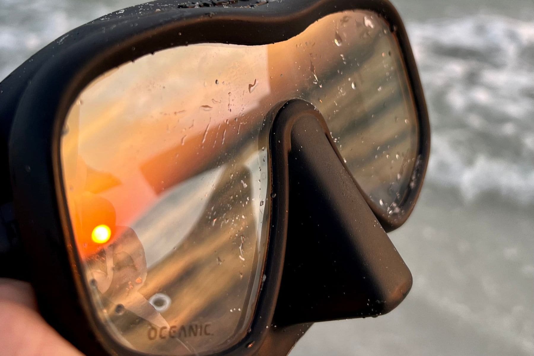 Oceanic Shadow single lens mask held up by the shore with water beading on the glass