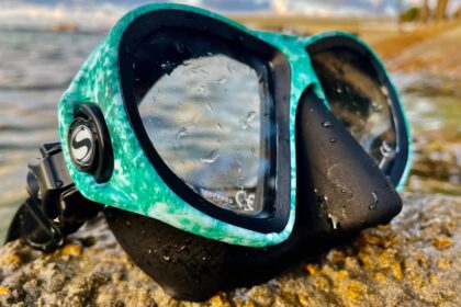 Sherwood Cruz mask in marbled teal, beaded with water on a rock at the shoreline