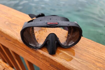 Hollis M1 frameless mask with a red logo, beaded with water on a wooden rail above the sea