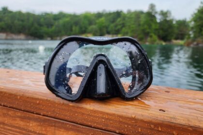 Atomic Aquatics Venom frameless mask with water beaded on its single lens, on a wooden rail by a lake