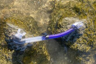 TUSA SP-0101 Hyperdry Elite II snorkel in purple and clear laid on a rock in shallow water