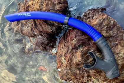 Blue Akona Jakarta snorkel with black mouthpiece resting on a weed-covered rock in shallow water