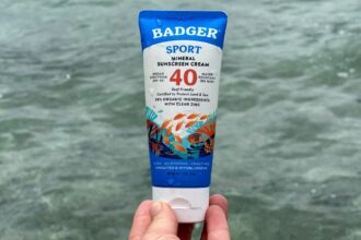 Hand holding a Badger Sport SPF 40 mineral sunscreen cream tube above clear green water