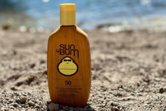 Sun Bum Original SPF 50 sunscreen lotion bottle standing in gravel with the sea behind