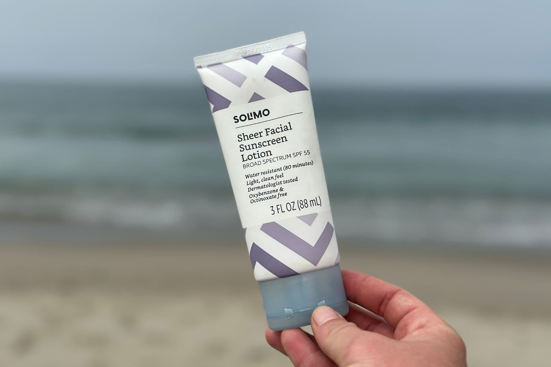 Hand holding an Amazon Solimo Sheer Facial Sunscreen Lotion SPF 55 tube on a beach