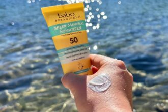 Hand holding a Babo Botanicals Sheer Mineral SPF 50 tube with a white smear of cream on the skin