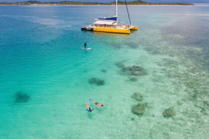 Two snorkelers over shallow coral in turquoise water with a yellow catamaran and paddleboarder behind
