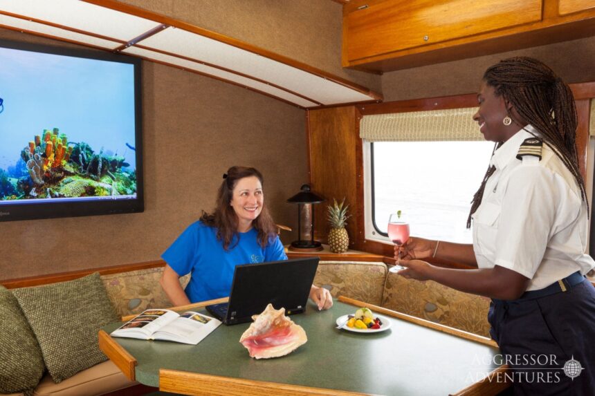 Guest reviewing dive log footage on a laptop while a crew member serves a drink