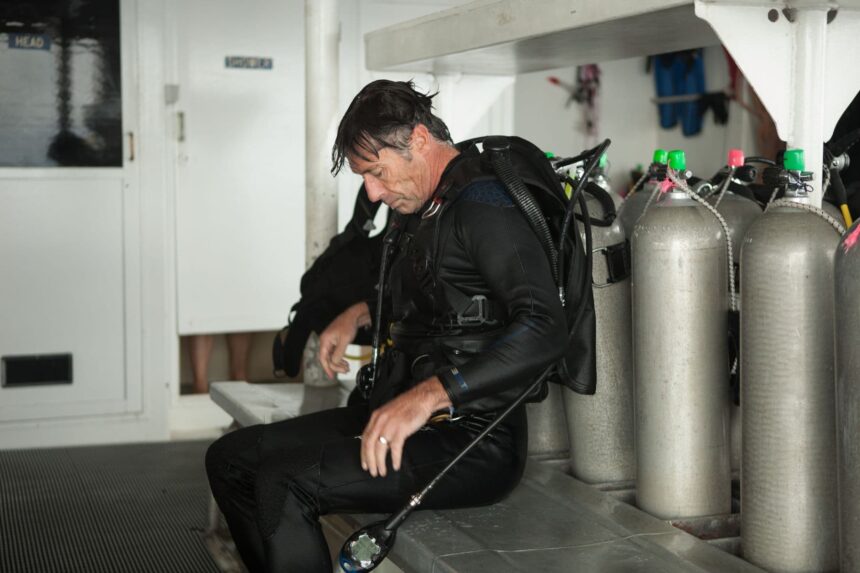 A diver seated on the dive deck adjusting gear beside a row of scuba tanks