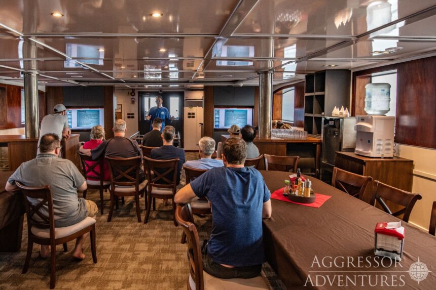 Guests seated in the Cocos Island Aggressor saloon watching a dive briefing on wall-mounted screens