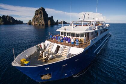 Blue Manta liveaboard crew waving from the bow with dramatic rock islands behind them