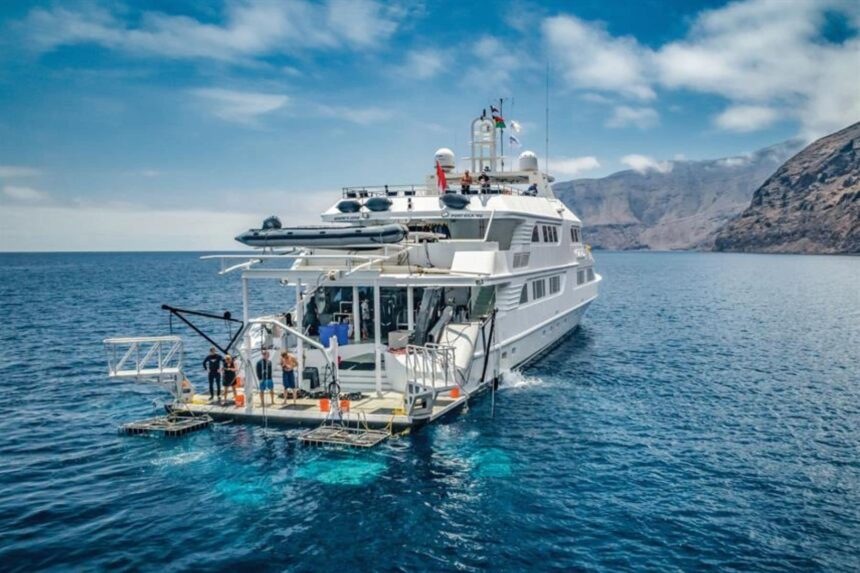 Nautilus Belle Amie liveaboard anchored near volcanic cliffs with divers on the dive platform