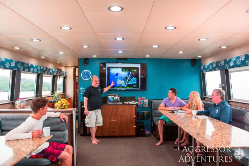 Guests gather around a wall-mounted screen in the Roatan Aggressor's lounge for a dive briefing