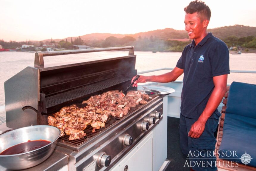 A crew member grills meat on deck as the sun sets over the harbor