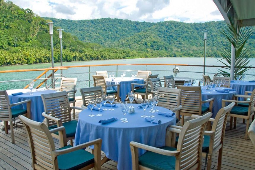 Tables set with blue linen on the Caledonian Sky open deck overlooking a forested shoreline