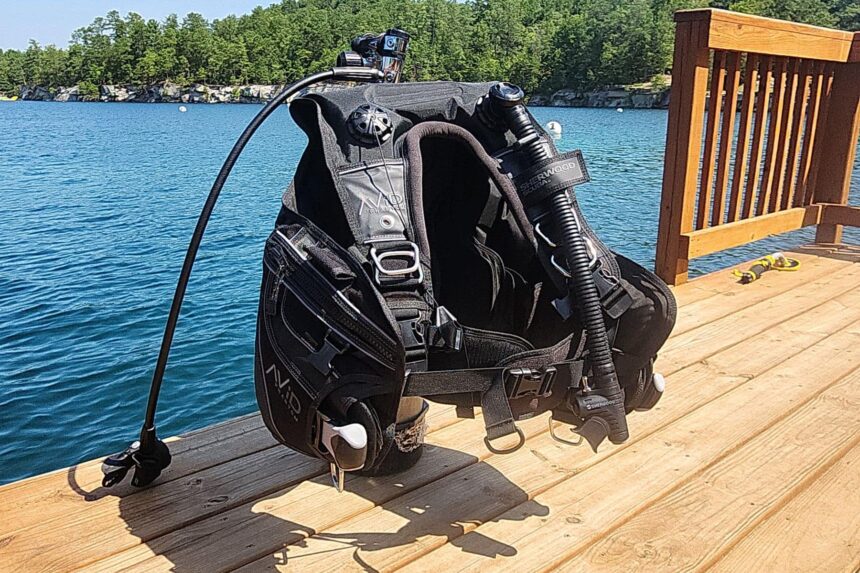 Sherwood Avid Phantom BCD standing on a dock beside a blue lake with the inflator hose extended