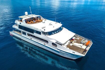 Solitude Adventurer liveaboard sailing with pale pink sails raised near a rocky shoreline