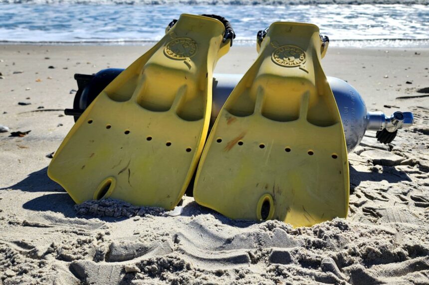 Yellow ScubaPro Jet Fins propped upright in wet sand with a dive cylinder behind them