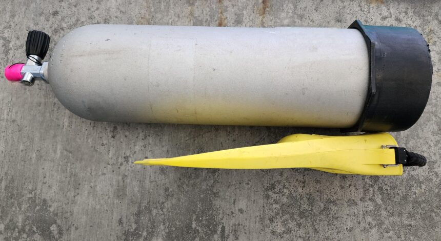 Yellow ScubaPro Jet Fin laid beside an aluminum 80 cylinder on concrete to compare length